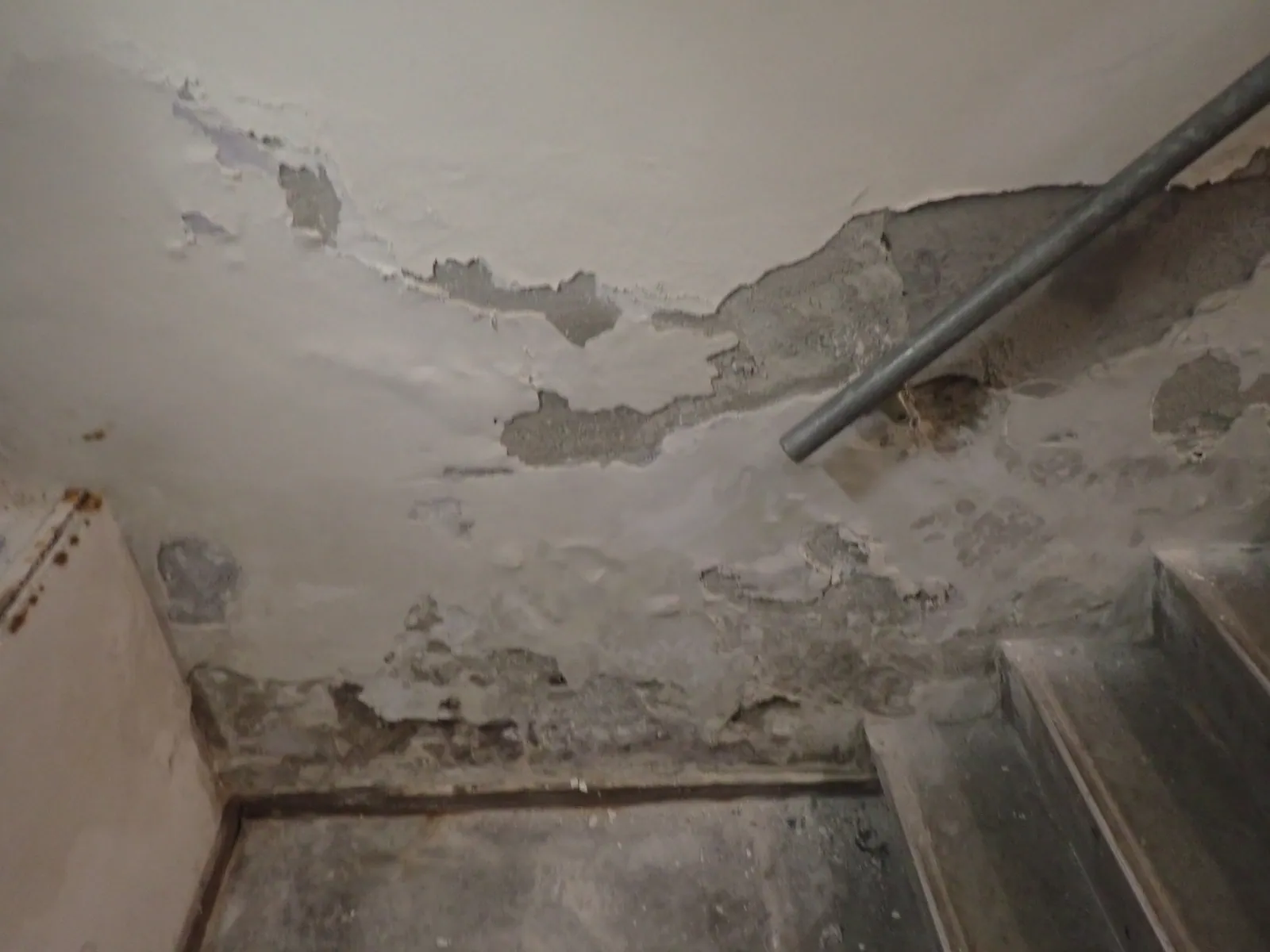 Kelderplafond met vochtschade, afgebrokkeld pleisterwerk rond leidingwerk en trap — beeld bij kelderonderzoek